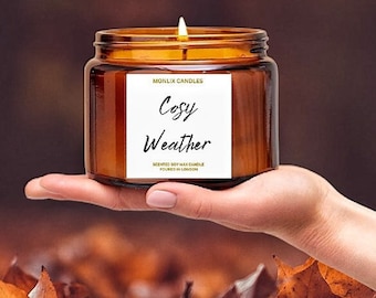 May include: A brown glass candle jar with a white label that reads "Monlix Candles Cozy Weather Scented Soy Wax Candle Poured in London". The candle is lit and the flame is visible. The candle is sitting on a bed of brown leaves.