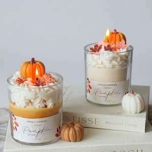 Puede incluir: Dos velas en tarro de cristal con aroma a especias de calabaza, cubiertas con cera blanca y una calabaza pequeña. Las velas están sobre una pila de libros con las palabras "MATISSE" y "VINCENT VAN GOGH" visibles. También hay calabazas decorativas adicionales.