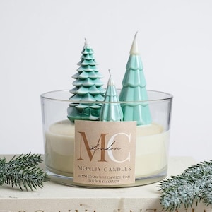 May include: A large glass candle with three teal tree-shaped candles inside. The candle has a cream-coloured wax and a label that reads "MONLIX CANDLES London". The candle is on a book with text and is decorated with faux evergreen branches.