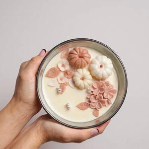 May include: A decorative candle in a clear glass container, featuring a cream-colored wax base. The candle is adorned with detailed, light-colored pumpkins, flowers, and leaves. The arrangement includes shades of pink, peach, and white, creating a seasonal, autumnal theme.