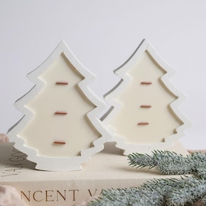 Peut inclure: Deux bougies blanches en forme de sapin avec un intérieur crème et trois accents marron. Les bougies sont posées sur un livre avec le texte "Vincent Van Gogh". Une branche de faux conifère ajoute une touche festive.