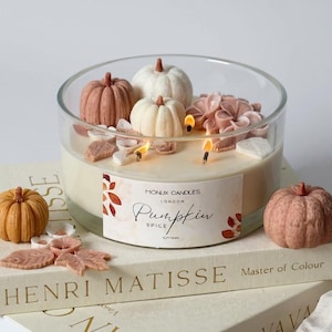 May include: A large glass candle with a pumpkin spice scent, decorated with miniature pumpkins and floral accents. The candle sits atop two books, with the text "Pumpkin Spice" visible on the label. The candle is lit, with three flames visible.