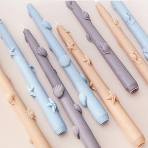 May include: A set of decorative candles with a nautical theme. The candles are in shades of blue, gray, and beige and feature seashells, starfish, and seahorses.