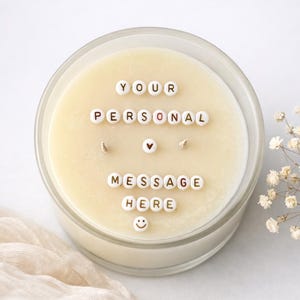 May include: A large, round candle in a clear glass container. The candle's surface is cream-colored, with the words "YOUR PERSONAL MESSAGE HERE" spelled out in white letter beads. A small heart and a smiley face are also included.