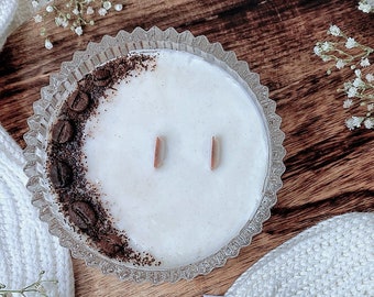 May include: A white candle with a coffee bean design on the side, in a clear glass jar with a decorative rim. The candle is sitting on a wooden surface with white knitted fabric.