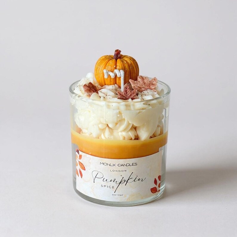 Puede incluir: Una vela de especias de calabaza en capas en un vaso transparente. La vela tiene una tapa en forma de calabaza con la letra "M", y est&aacute; decorada con glaseado blanco y hojas de oto&ntilde;o. La etiqueta dice "MONEX CANDLES LONDON Pumpkin Spice NEW YORK".
