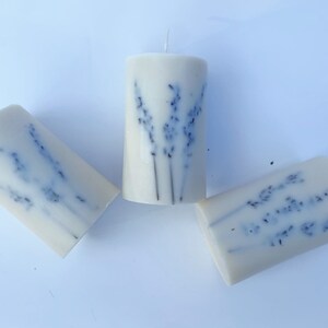 May include: Three white pillar candles with dried lavender flowers embedded in the wax. The candles are arranged in a triangular pattern on a white surface.