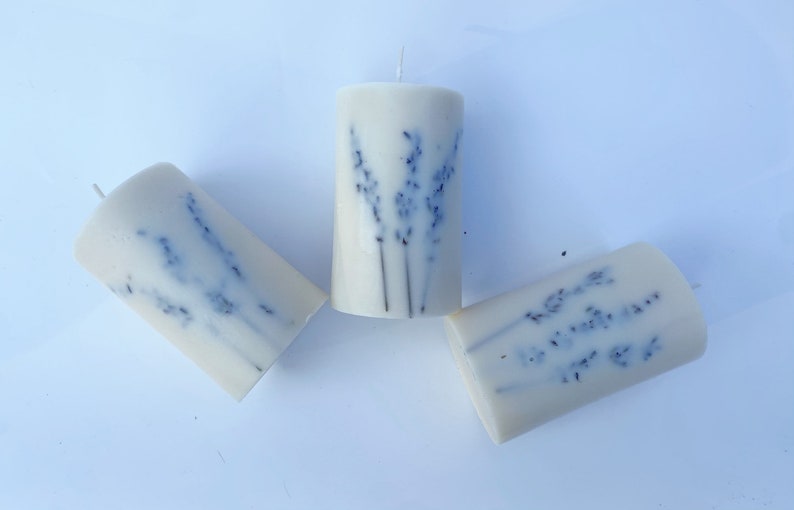 May include: Three white pillar candles with dried lavender flowers embedded in the wax. The candles are arranged in a triangular pattern on a white surface.