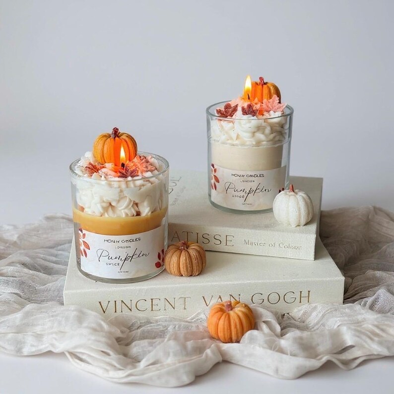 Puede incluir: Dos velas en tarro de cristal con aroma a especias de calabaza, cubiertas con cera blanca y una calabaza peque&ntilde;a. Las velas est&aacute;n sobre una pila de libros con las palabras "MATISSE" y "VINCENT VAN GOGH" visibles. Tambi&eacute;n hay calabazas decorativas adicionales.