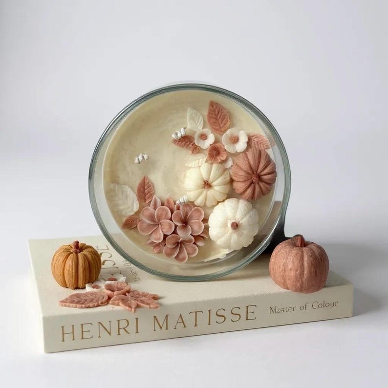 May include: A decorative glass bowl with a cream-colored interior, featuring sculpted pumpkins and flowers in shades of white, peach, and brown. The bowl sits atop a book titled "HENRI MATISSE Master of Colour", with additional pumpkin and leaf decorations.