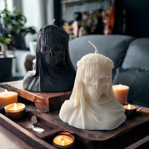 May include: Two decorative candles in the shape of a person's head wearing a veil. The candles are on a wooden tray with other candles.