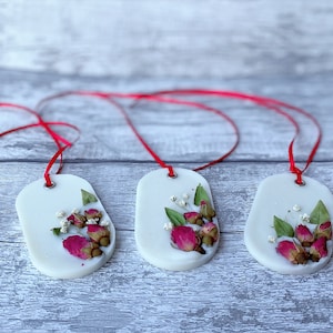 May include: Three white oval wax melts with red ribbon hangers. Each melt features pressed dried flowers, including pink roses and white baby's breath.