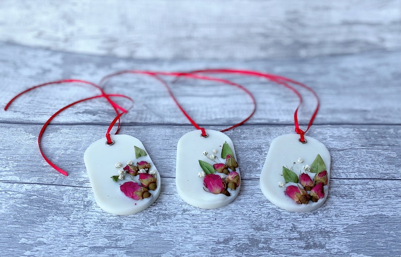 May include: Three white oval wax melts with red ribbon hangers. Each melt features pressed dried flowers, including pink roses and white baby's breath.