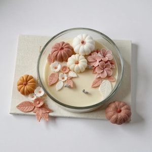 May include: A clear glass candle holder filled with cream-colored wax, decorated with small pumpkins, flowers, and leaves in shades of orange, pink, and white. The candle sits on a neutral-toned rectangular surface.