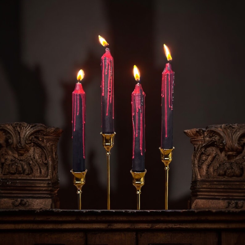 May include: Five black candles with red wax dripping down the sides are lit and placed on gold candle holders. The candles are arranged in a row on a dark wooden surface.