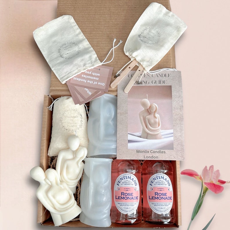 May include: A gift box set for couples with two white ceramic candles, two bottles of rose lemonade, two white fabric bags, and a guide for making the candles. The box is open and the items are visible.