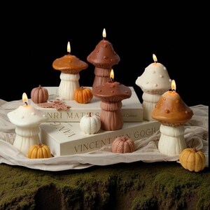 May include: A collection of mushroom-shaped candles in various shades of brown and white, with lit wicks. Small pumpkin-shaped decorations are scattered around the candles. The candles are arranged on books and a white cloth, set against a dark background.