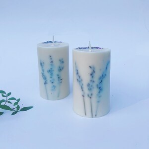 May include: Two white pillar candles with a lavender design. The candles have a light blue and purple floral pattern.