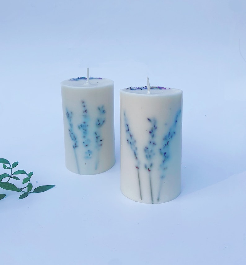 May include: Two white pillar candles with a lavender design. The candles have a light blue and purple floral pattern.