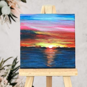 May include: A small painting on canvas depicting a sunset over a body of water. The sky is a vibrant mix of pink, orange, and yellow, with a few clouds. The water is a deep blue, reflecting the colours of the sky. The painting is on a small wooden easel.