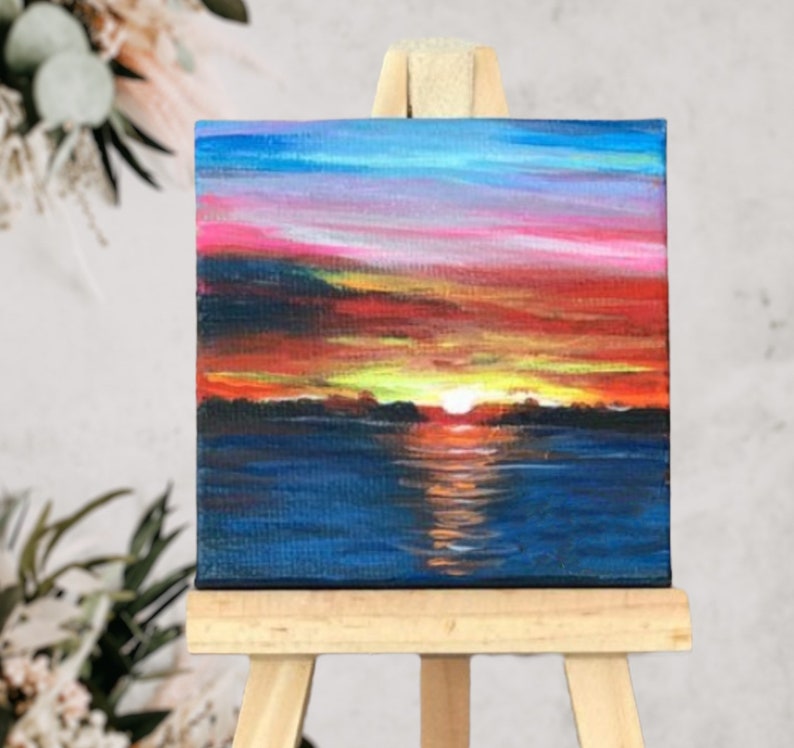 May include: A small painting on canvas depicting a sunset over a body of water. The sky is a vibrant mix of pink, orange, and yellow, with a few clouds. The water is a deep blue, reflecting the colours of the sky. The painting is on a small wooden easel.