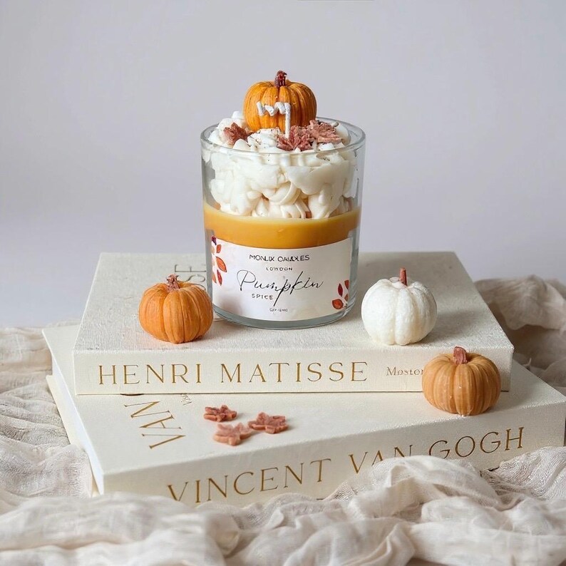 Puede incluir: Una vela de cristal transparente con aroma a especias de calabaza, cubierta con cera blanca y una peque&ntilde;a calabaza naranja. La vela est&aacute; sobre una pila de libros con calabazas decorativas. La etiqueta de la vela dice "Pumpkin Spice".