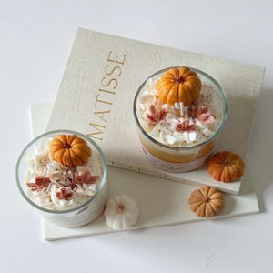 Puede incluir: Dos velas de vidrio con cera tipo crema batida, adornadas con decoraciones de calabazas naranjas y detalles oto&ntilde;ales. Las velas est&aacute;n sobre una superficie blanca con la palabra "MATISSE". Decoraciones de calabaza adicionales est&aacute;n cerca.
