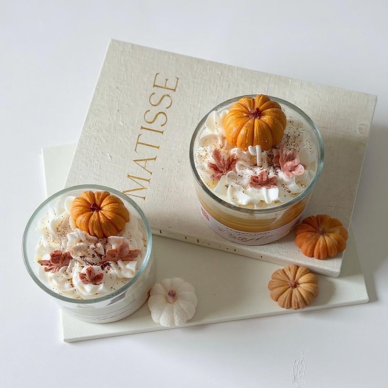 Puede incluir: Dos velas de vidrio con cera tipo crema batida, adornadas con decoraciones de calabazas naranjas y detalles oto&ntilde;ales. Las velas est&aacute;n sobre una superficie blanca con la palabra "MATISSE". Decoraciones de calabaza adicionales est&aacute;n cerca.