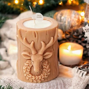 May include: A tan-colored candle shaped like a tree stump, featuring a deer head with antlers and a wreath. The candle has a white interior and a wick. The label on the wick says "Montix Candles". A lit candle and Christmas lights are in the background.