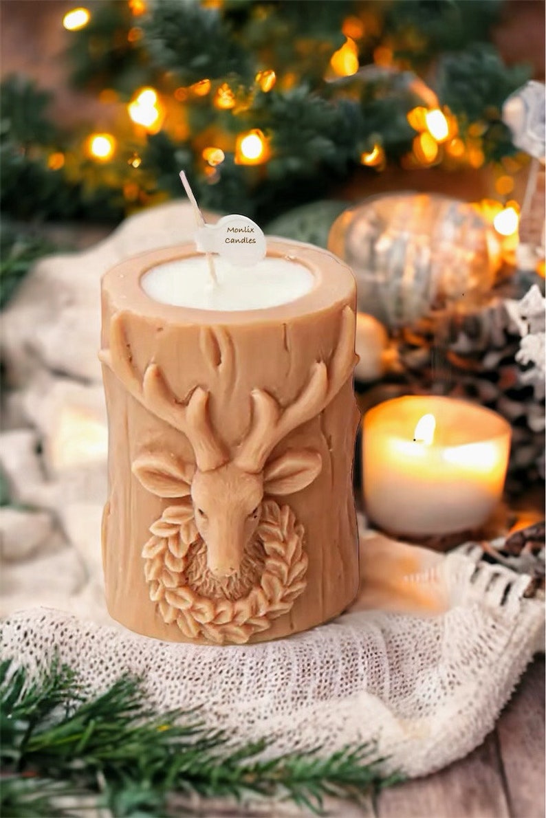 May include: A tan-colored candle shaped like a tree stump, featuring a deer head with antlers and a wreath. The candle has a white interior and a wick. The label on the wick says "Montix Candles". A lit candle and Christmas lights are in the background.