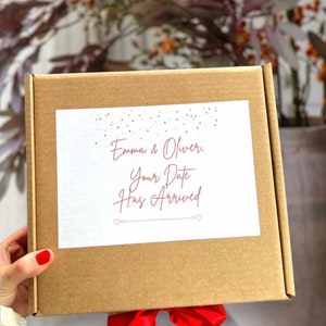 May include: A brown cardboard box with a red ribbon tied around it. The box has a white label with the text "Emma & Oliver, Your Date Has Arrived" in red cursive writing.