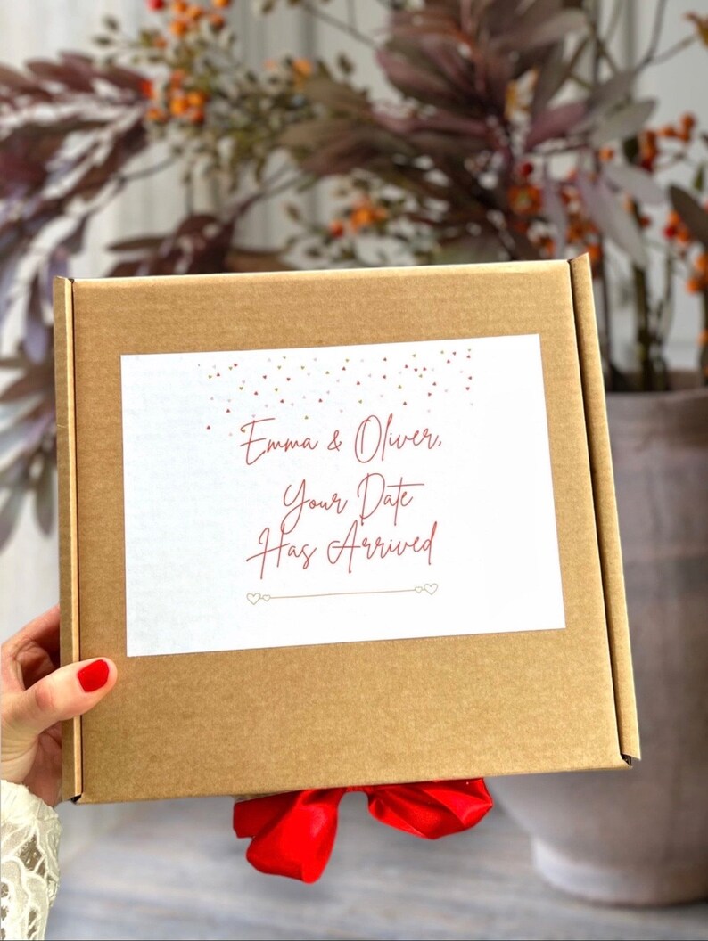 May include: A brown cardboard box with a red ribbon tied around it. The box has a white label with the text "Emma & Oliver, Your Date Has Arrived" in red cursive writing.