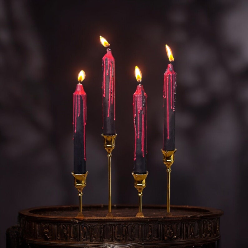 May include: Four black candles with red wax dripping down the sides are lit and placed on gold candle holders on a dark wooden surface.