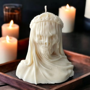 May include: A white candle shaped like a skull wearing a veil. The candle is sitting on a wooden tray.