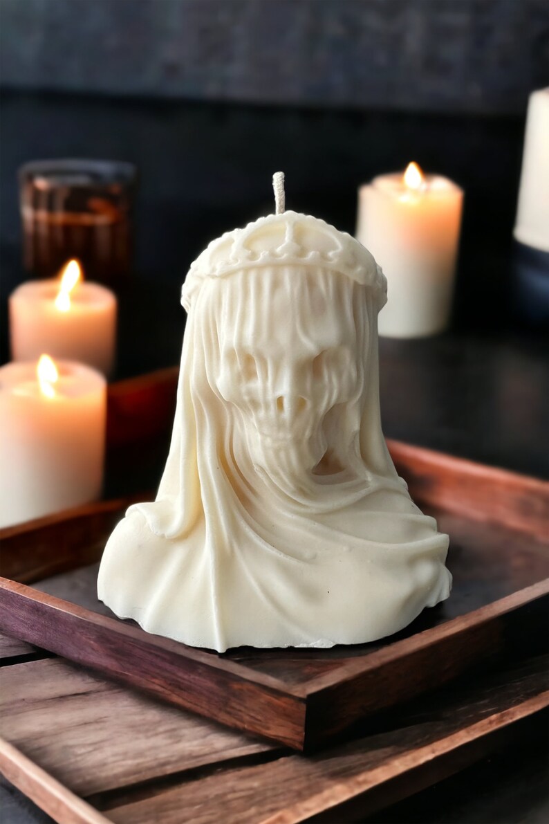 May include: A white candle shaped like a skull wearing a veil. The candle is sitting on a wooden tray.
