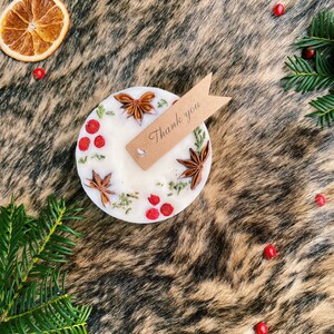 May include: A white candle with a brown tag that says "Thank you". The candle is decorated with dried berries, star anise, and greenery.