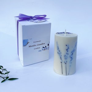 May include: A white candle with dried lavender flowers embedded in the wax. The candle is in a white box with a purple ribbon and a label that says "Handmade Monlix Candles London".