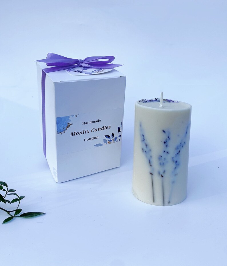 May include: A white candle with dried lavender flowers embedded in the wax. The candle is in a white box with a purple ribbon and a label that says "Handmade Monlix Candles London".