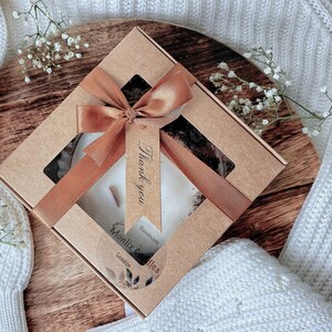 May include: A brown cardboard box with a clear window showing a white candle with a brown ribbon and a tag that says "Thank you". The box is on a wooden surface and has a label that says "Monlix Candles" and "Handmade in London".
