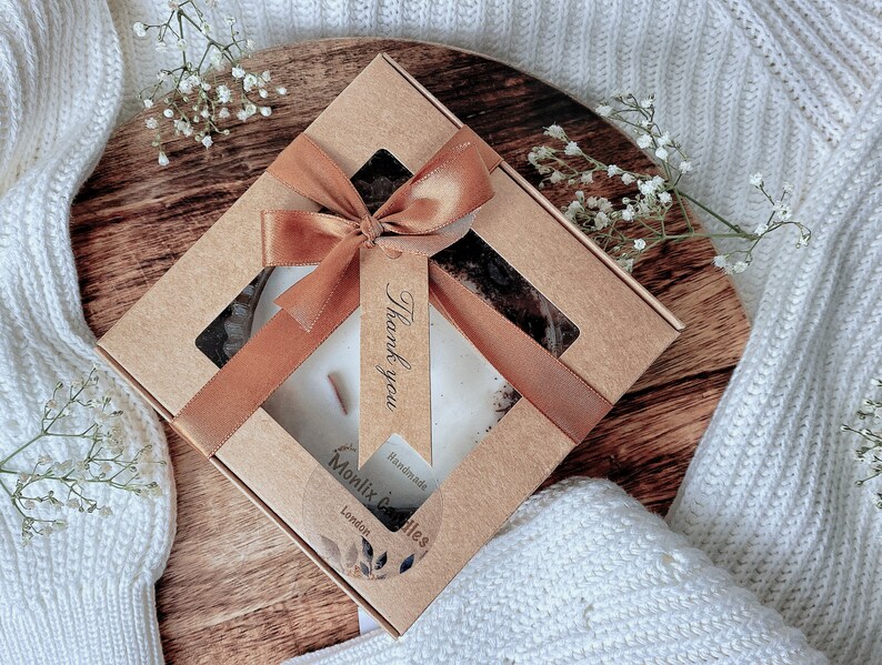 May include: A brown cardboard box with a clear window showing a white candle with a brown ribbon and a tag that says "Thank you". The box is on a wooden surface and has a label that says "Monlix Candles" and "Handmade in London".