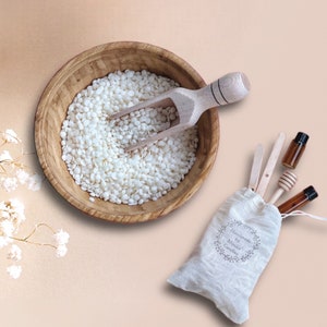 May include: A wooden bowl filled with white wax flakes, a wooden scoop, and two small glass bottles with droppers. A small white bag with the text "Handmade by Moxie Candles" is in the lower right corner.