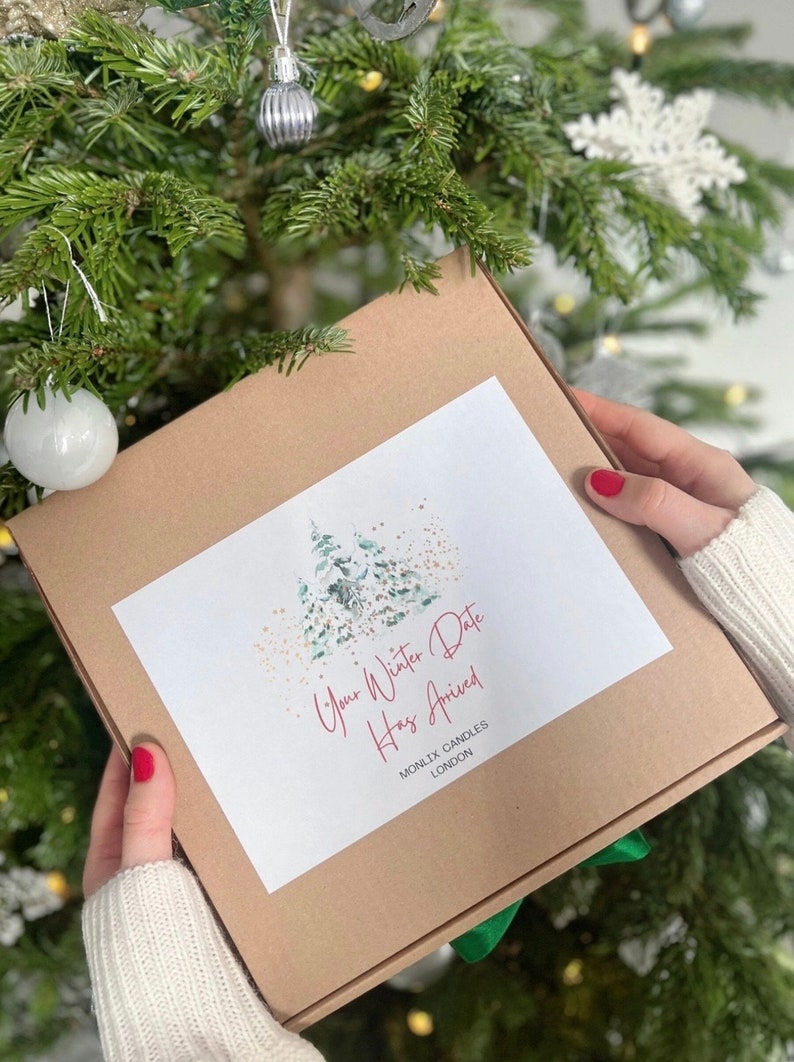 May include: A brown box with a white label that says "Your Winter Date Has Arrived" and "MONLIX CANDLES LONDON". The label also features a watercolor illustration of a forest scene with green trees and gold glitter.