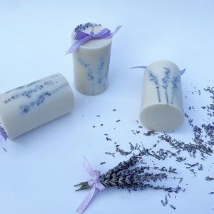 May include: Three white pillar candles with lavender flowers embedded in the wax. The candles are tied with purple ribbon and are surrounded by dried lavender flowers.