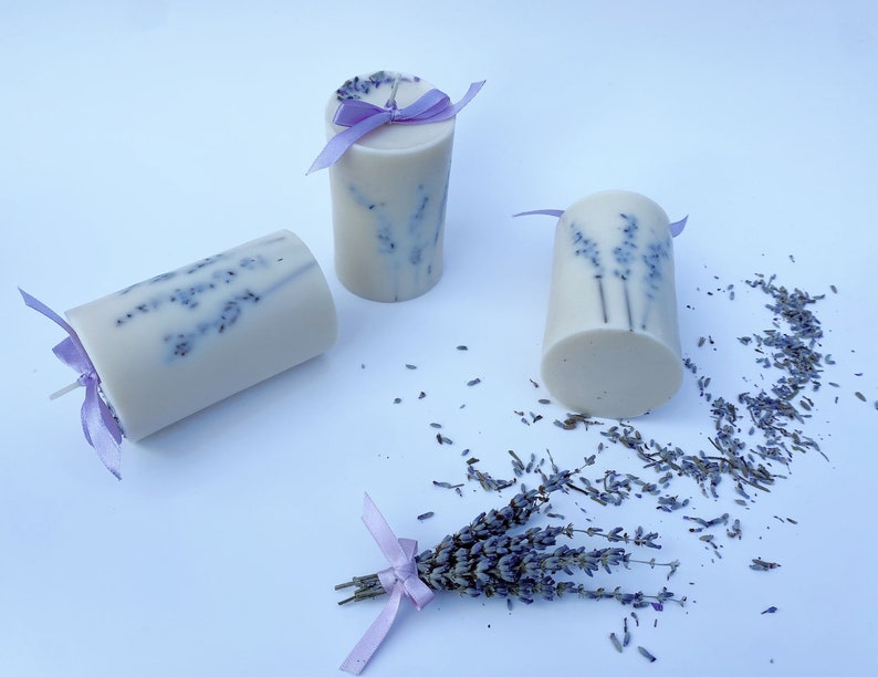 May include: Three white pillar candles with lavender flowers embedded in the wax. The candles are tied with purple ribbon and are surrounded by dried lavender flowers.