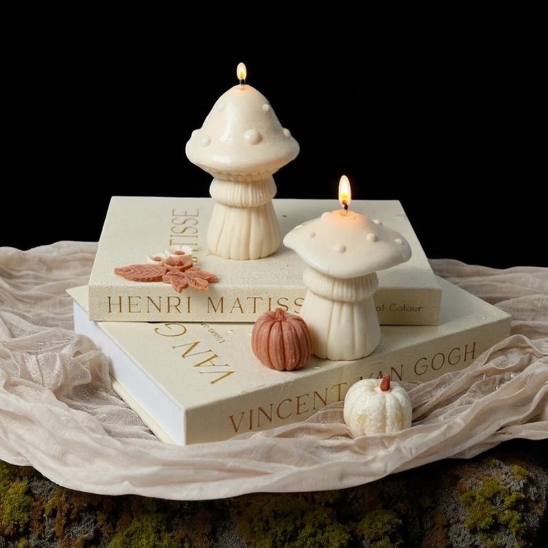 May include: Two lit mushroom-shaped candles with a textured surface sit atop books with gold lettering. A small pumpkin and leaf accents are placed nearby. The candles are ivory and the pumpkins are shades of orange and white.