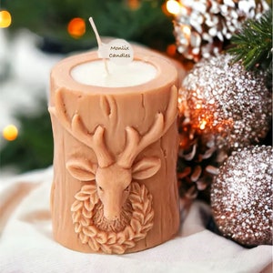 May include: A tan-colored candle shaped like a tree trunk, featuring a carved deer head with antlers and a wreath. The candle has a white wax interior and a wick. The text "Monlix Candles" is on a small tag. Festive decor.