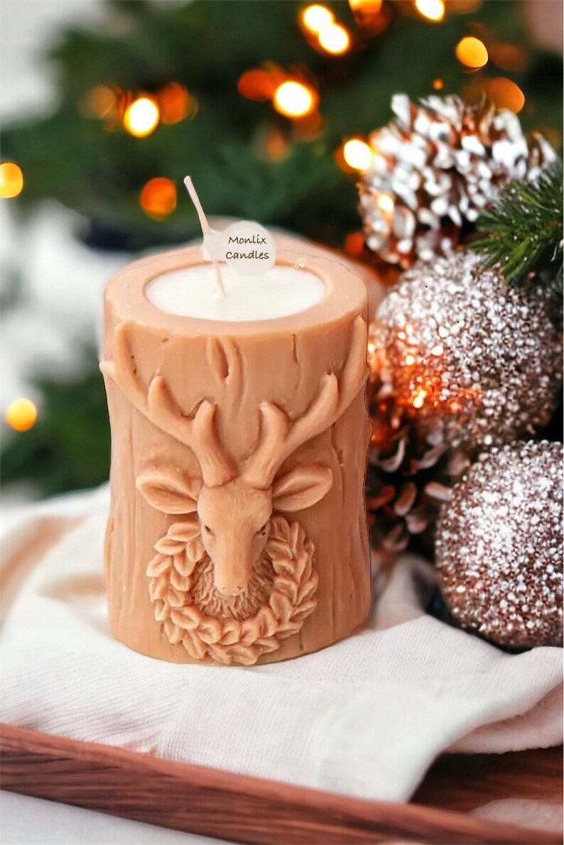 May include: A tan-colored candle shaped like a tree trunk, featuring a carved deer head with antlers and a wreath. The candle has a white wax interior and a wick. The text "Monlix Candles" is on a small tag. Festive decor.