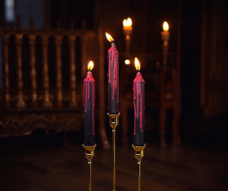 May include: Three black candles with red wax dripping down the sides are lit and placed on gold candle holders. The candles are in front of a dark wooden background.