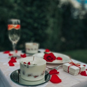May include: A white candle with red rose petals embedded in it sits on a mirrored plate. The candle is surrounded by rose petals on a white tablecloth. A small white gift box with a red ribbon is in the background.