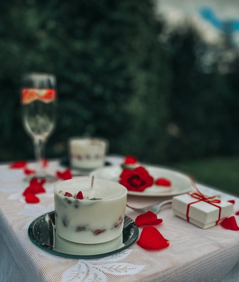 May include: A white candle with red rose petals embedded in it sits on a mirrored plate. The candle is surrounded by rose petals on a white tablecloth. A small white gift box with a red ribbon is in the background.